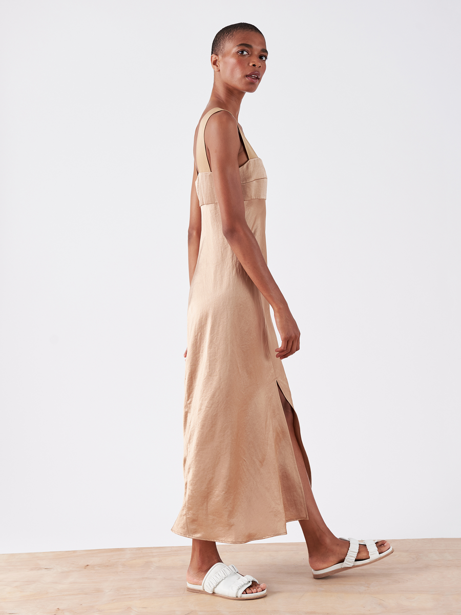 A person models the stylish Bias Oona Dress - Final Sale by Zero + Maria Cornejo, standing sideways in the sleeveless, light brown linen-blend maxi with a square neckline and side slit. White sandals complete the look against a wooden surface and plain white background.