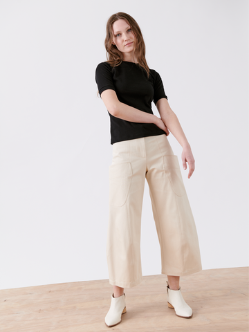 A person with long hair is standing against a plain white background. They are wearing a black short-sleeve top, Curved Eda Culotte from Zero + Maria Cornejo, and white ankle boots. Their hands are relaxed by their sides. The floor is light-colored wood.