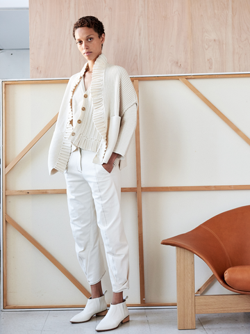 Person with short hair stands in a relaxed pose against a geometric framed backdrop in New York. They're dressed in a white ensemble: a chunky knit cardigan, ribbed tank top, Zero + Maria Cornejo Slim Takeo Pant, and matching ankle boots. An orange-brown chair is partially visible.