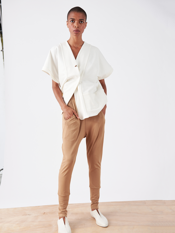 A person confidently stands on a wooden floor against a white backdrop, wearing a white wrap-style short-sleeve top and Zero + Maria Cornejo's tan Drop Gabi Pant - Final Sale with an elastic waist. White slip-on shoes complete the stylish, poised look.