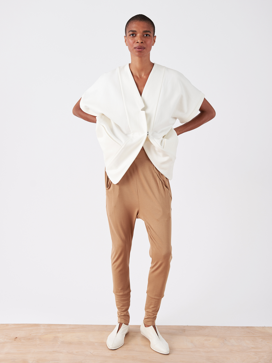 A person models the Drop Gabi Pant - Final Sale by Zero + Maria Cornejo against a plain white backdrop. The tan trousers, with an elastic waist for comfort, are paired with a loose white top and white shoes. They have short hair.