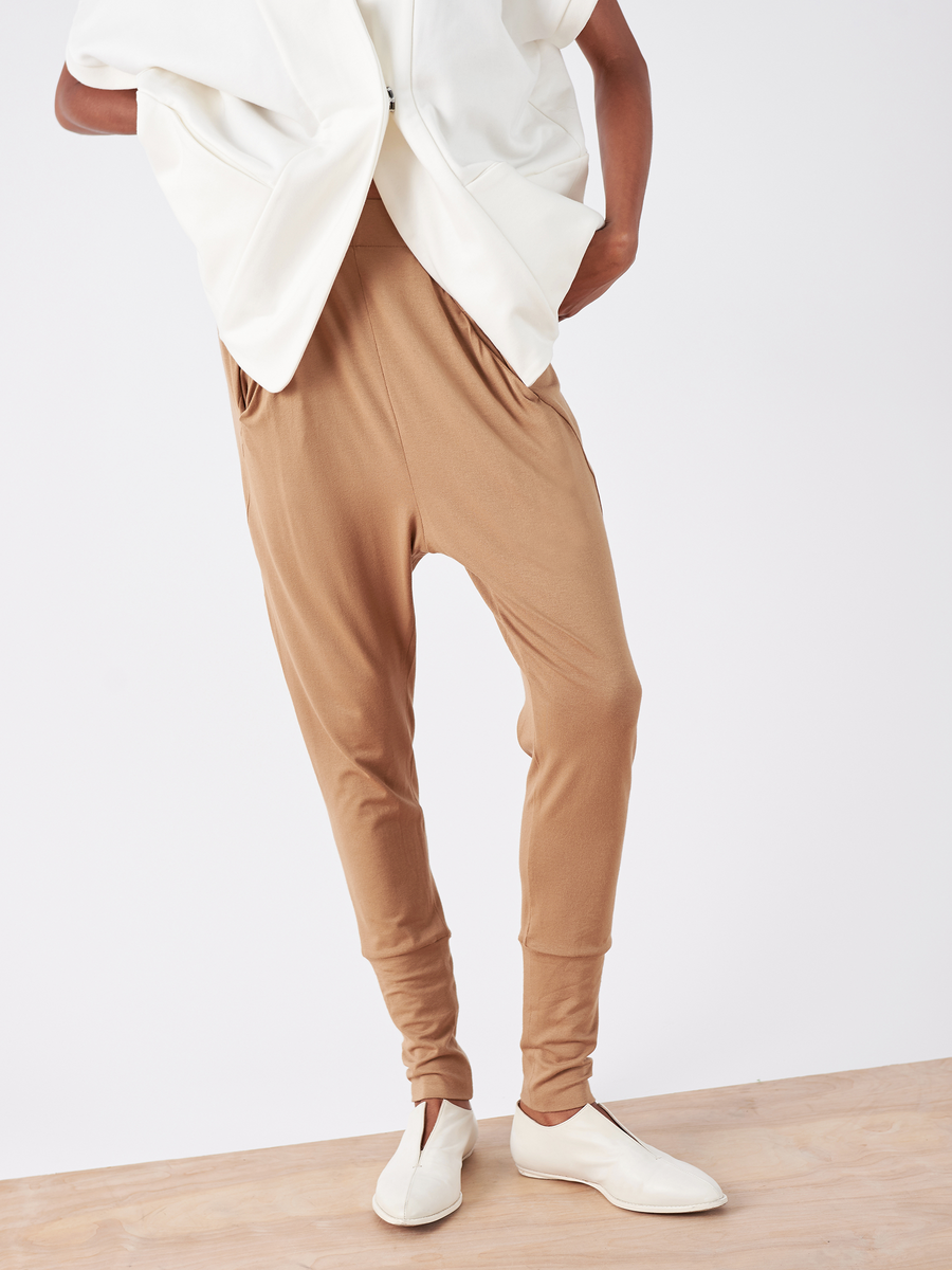 A person in a white top and the tan Drop Gabi Pant by Zero + Maria Cornejo, featuring an elastic waist, stands on a light wooden floor with hands in pockets and off-white shoes. The image is cropped above the shoulders, showcasing a blend of comfort and modernity.