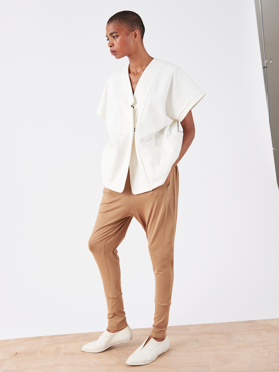 Against a white background, this person wears a loose-fitting, short-sleeved white top with Zero + Maria Cornejo's Drop Gabi Pant in tan viscose, showcasing an elastic waist and drop-crotch design. Beige slip-on shoes complete the look. The individual has short hair and gazes to the side.