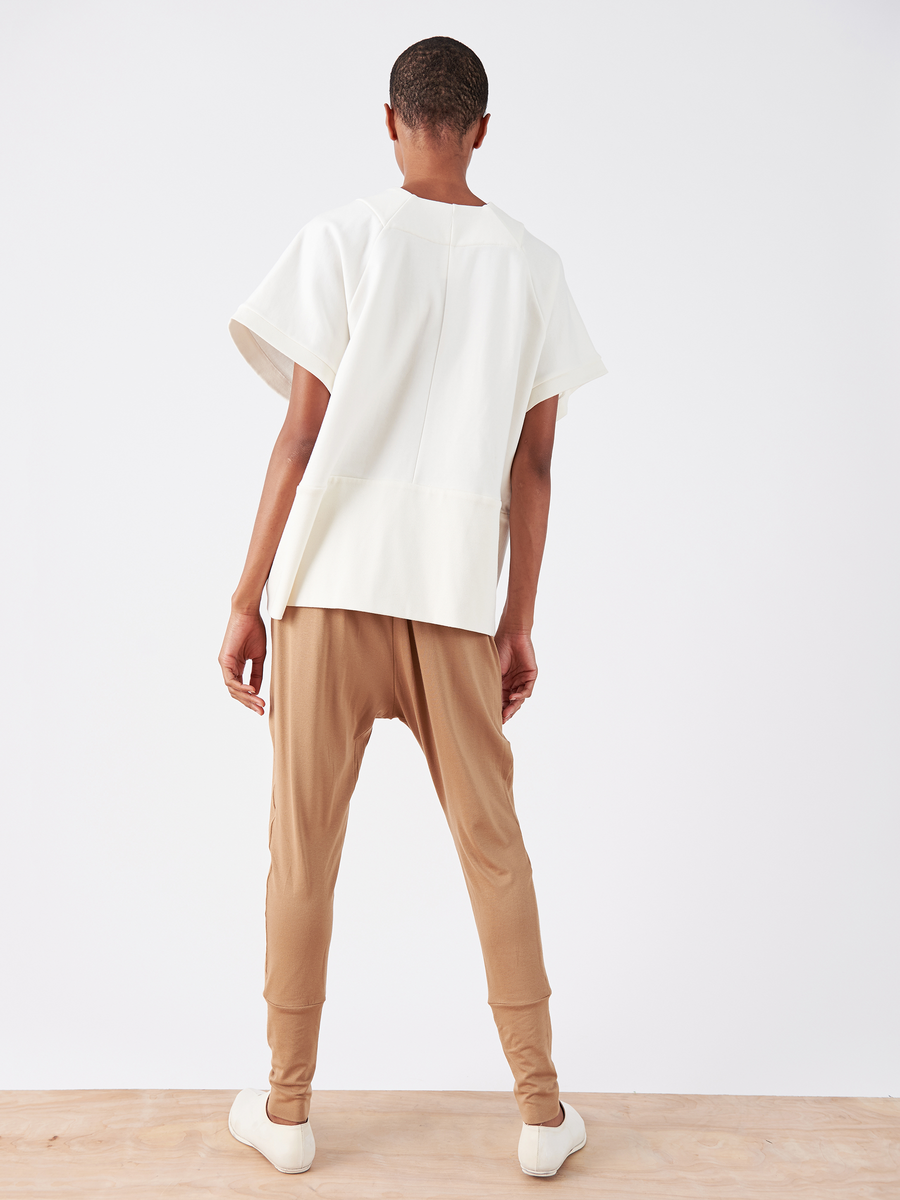 A person is on a wood floor with a white background, wearing a white short-sleeve top and tan Drop Gabi Pant by Zero + Maria Cornejo, featuring an elastic waist. The outfit's back and their white shoes are showcased.