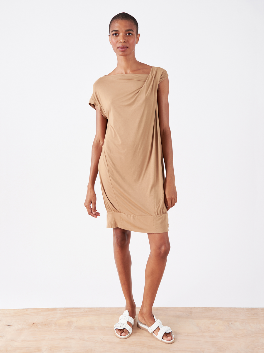 Standing on a wooden floor, the person wears the Twist T Dress - Final Sale by Zero + Maria Cornejo, a light brown knee-length dress with an asymmetrical neckline, paired with white sandals against a plain white background.