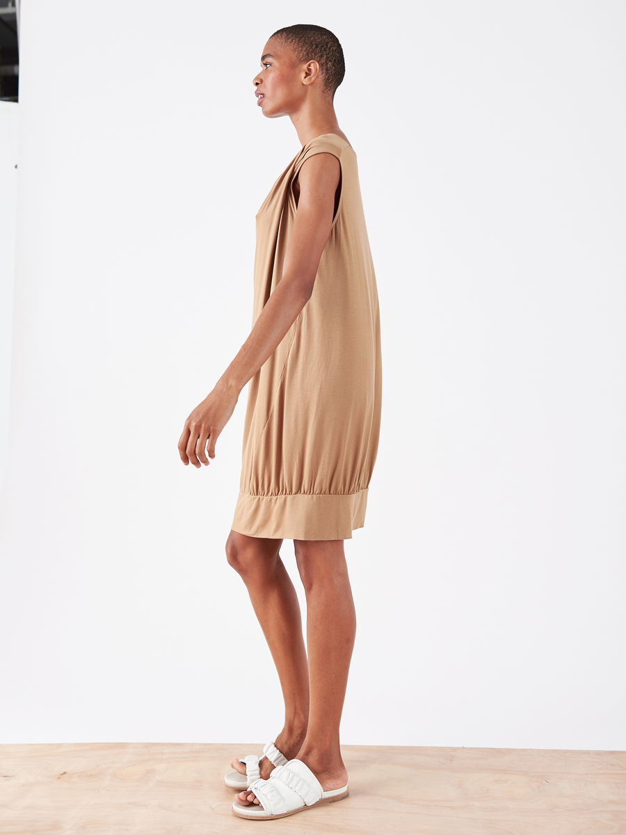 A model stands in profile against a white backdrop, wearing the Twist T Dress by Zero + Maria Cornejo. This loose-fitting tan dress features an asymmetrical neckline and knee-length hem, complemented by white slide sandals, and is made from biodegradable material for eco-friendliness.