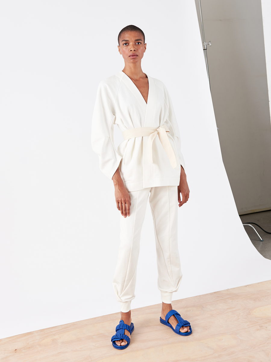 In a minimalist studio with a plain white backdrop, a person wears Zero + Maria Cornejo's Akeo Sweatpant - Final Sale paired with a French Terry kimono-style top and blue sandals, highlighting the outfit's elegance and comfort with an elastic banded waist.