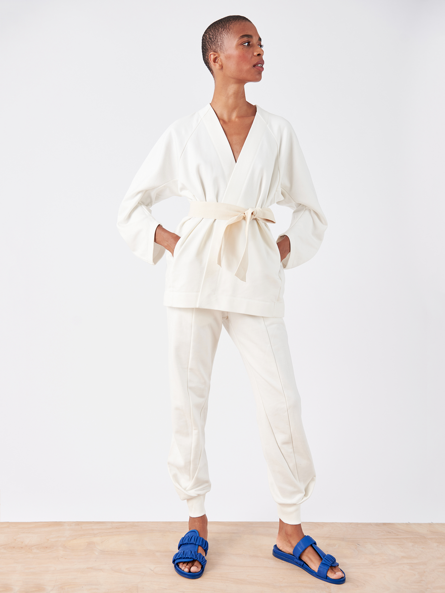 A person confidently poses in the chic Akeo Sweatpant with an elastic waist by Zero + Maria Cornejo, paired with a white belted top and blue sandals. One hand rests in a pocket while the other is on their hip, all set against a plain white background that highlights the stylish ensemble.