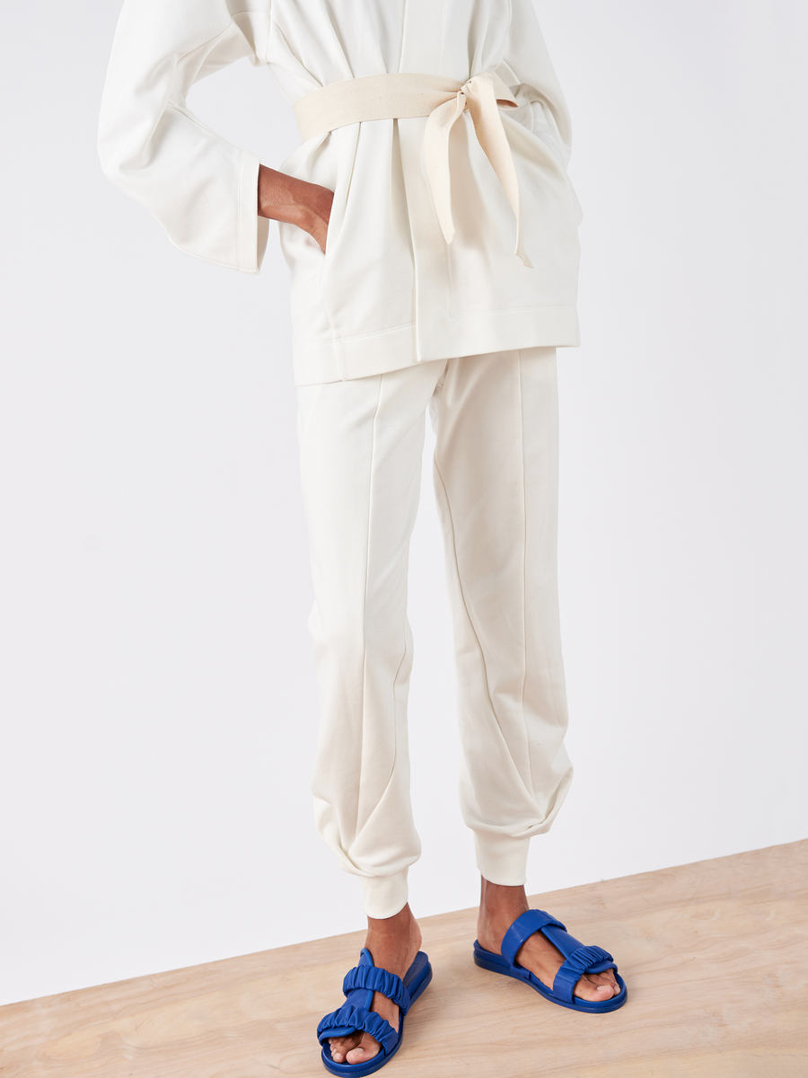 Standing on a wooden floor in an off-white ensemble, featuring the Akeo Sweatpant by Zero + Maria Cornejo, with French Terry top and cuffed pants showcasing an elastic waist. Their blue sandals have ruffled straps and contrast against a simple white background.