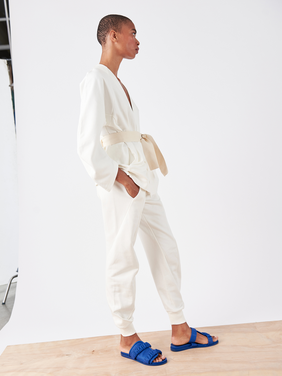 A person stands on a wooden platform against a white background, wearing the Akeo Sweatpant by Zero + Maria Cornejo, paired with a white jumpsuit, beige belt, and blue slide sandals. The ensemble offers classic French Terry comfort and style while they gaze to the side.