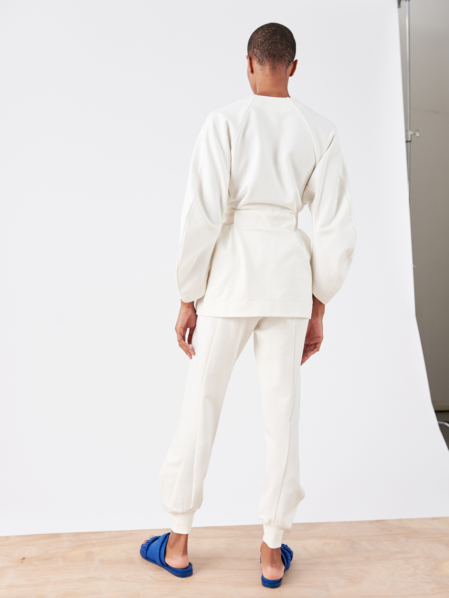 A person stands on a light wood floor against a white backdrop, facing away. They're wearing an all-white outfit from Zero + Maria Cornejo, including the Akeo Sweatpant made of soft French Terry fabric with elastic banded waist pants and loose sleeves, paired with blue sandals.