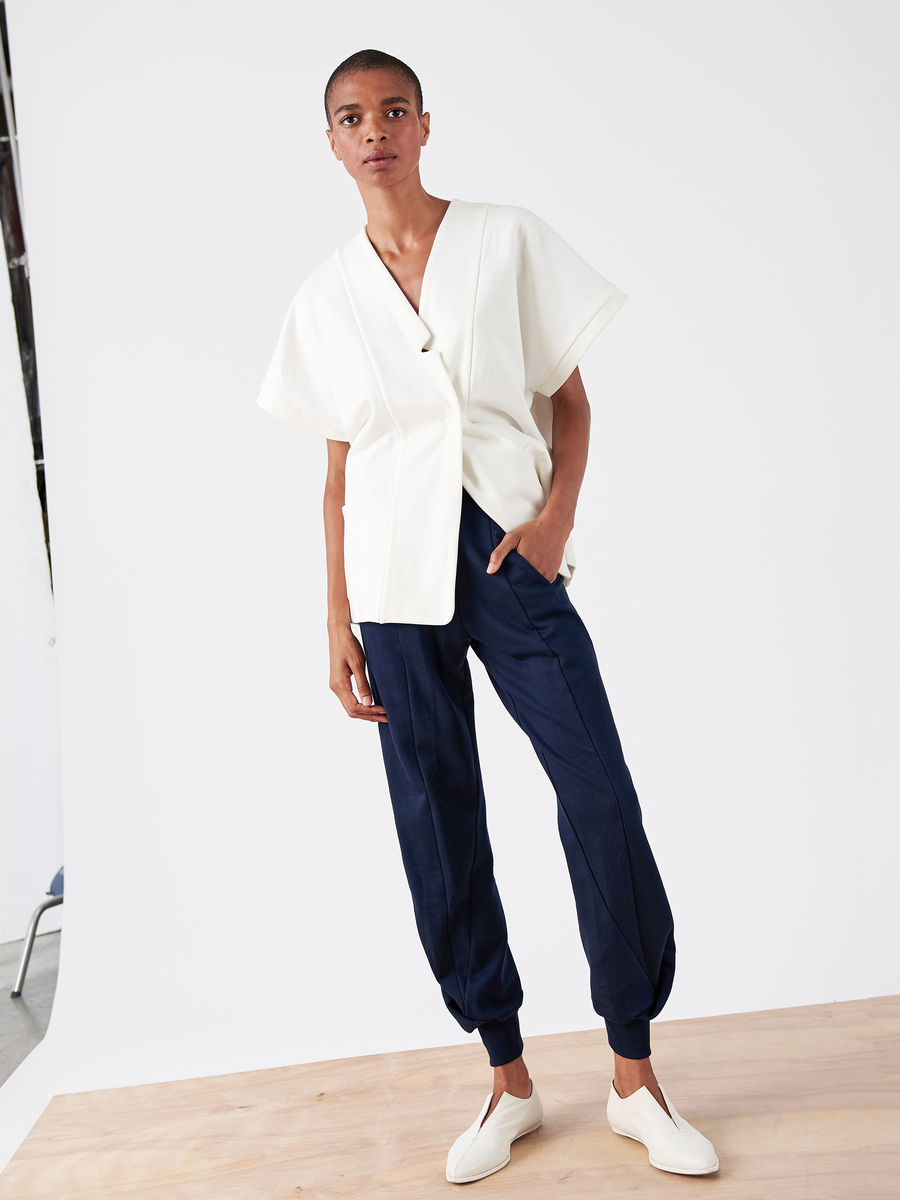 A person wearing a white V-neck top with short sleeves and Zero + Maria Cornejo's dark blue Akeo Sweatpant - Final Sale, featuring an elastic banded waist, stands on a light wooden platform. They have short hair and wear white slip-on shoes, posing against a plain white background.