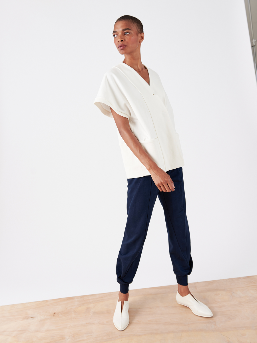 A person, clad in a loose-fitting white V-neck top and Zero + Maria Cornejo's Akeo Sweatpant - Final Sale, stands on a wooden floor against a plain white background. White slip-on shoes complete the look as they strike a pose with hands in pockets and slight lean.