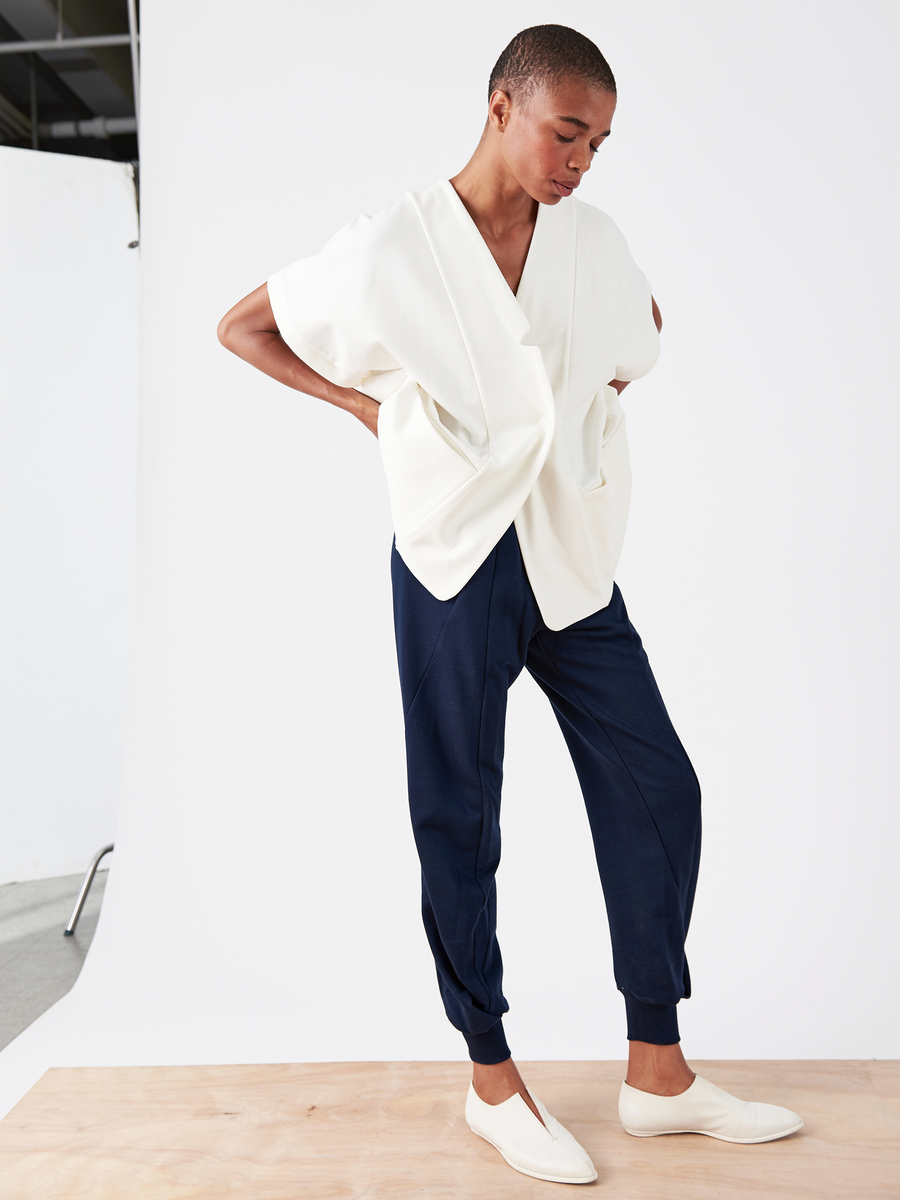 Against a white background, a person poses on a wooden platform wearing an oversized white top with wide sleeves, navy blue Akeo Sweatpants by Zero + Maria Cornejo, and white slip-on shoes. They pose with bent arms while gazing down to the side.