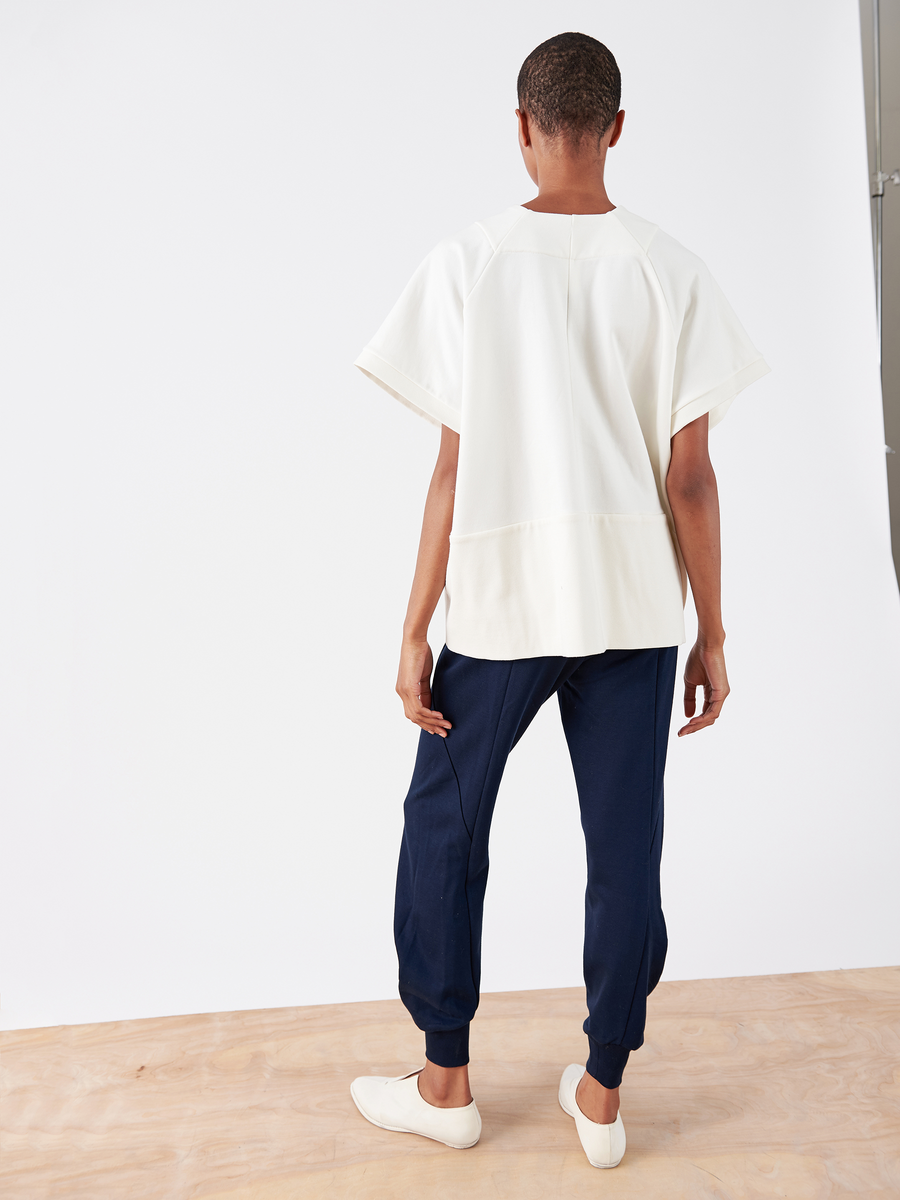 Against a white wall on a wooden floor, a person faces away, wearing a loose white short-sleeved top and navy Akeo Sweatpants by Zero + Maria Cornejo with an elastic waist. White slip-on shoes complete the outfit.