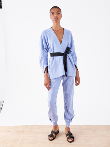 A person in Zero + Maria Cornejo's Akeo Sweatpant stands on a wooden platform against a white background. They're dressed in a light blue French Terry outfit with a kimono-style top, black belt, and black sandals. The pants have an elastic waist, and the person maintains a neutral expression while looking forward.