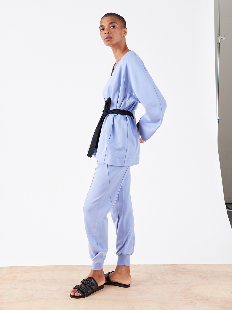 On a wooden floor, someone wears the Akeo Sweatpant by Zero + Maria Cornejo. The light blue outfit features a black belt and sandals, reminiscent of French Terry fabric, exuding comfort and style as they face slightly sideways against a plain white background.