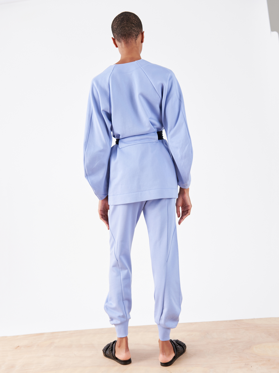 On a wooden surface, someone faces away wearing Zero + Maria Cornejo's Akeo Sweatpant—a cozy French terry set in light blue with an elastic waist, accented by a belt and paired with black slide sandals.