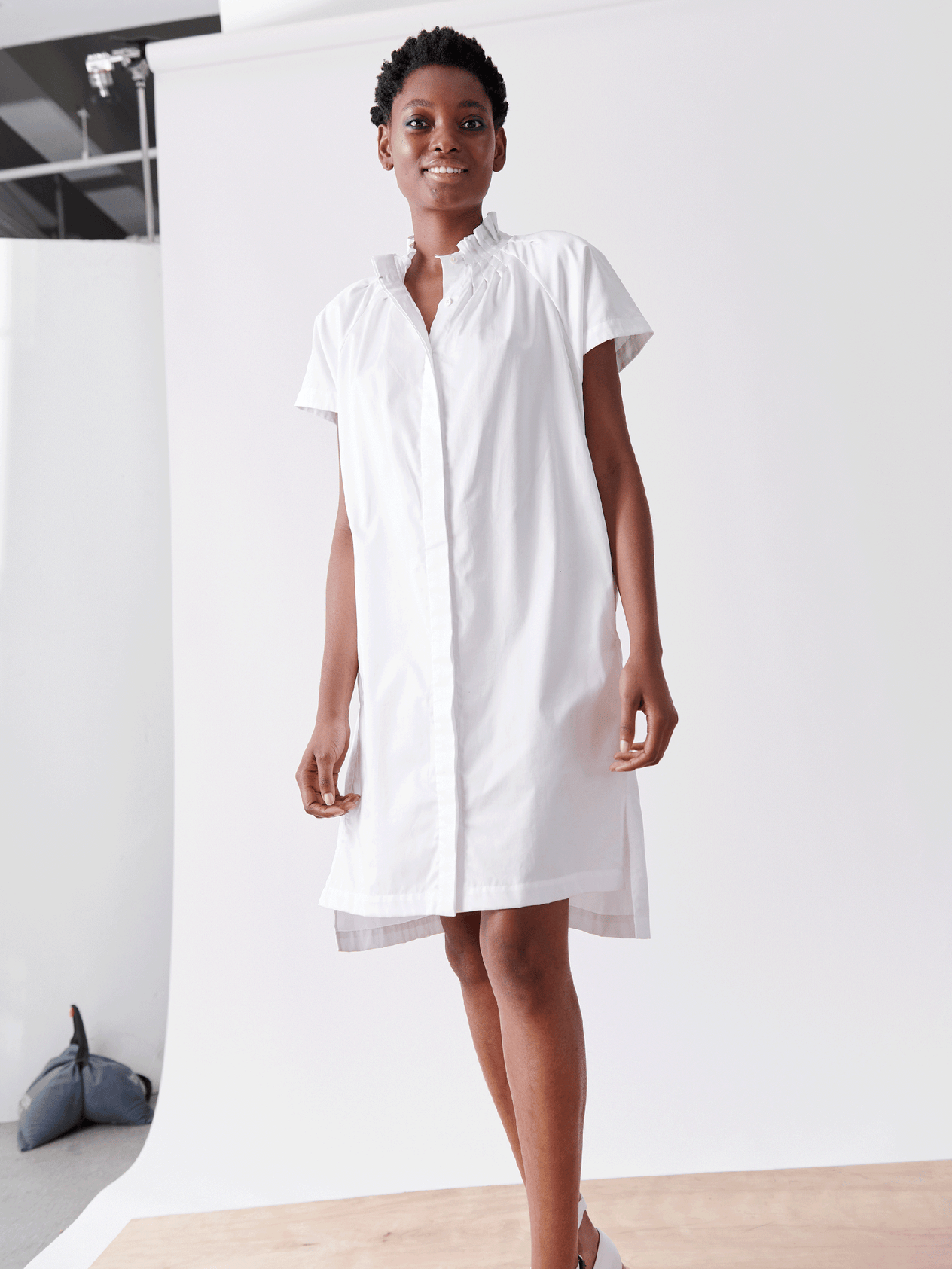 A person wearing a short-sleeved knee-length Pintuck Day Dress by Zero + Maria Cornejo stands on a wooden platform against a plain white and light gray background. They have short hair and are smiling confidently. A blue bag is seen on the left side, placed on the floor.