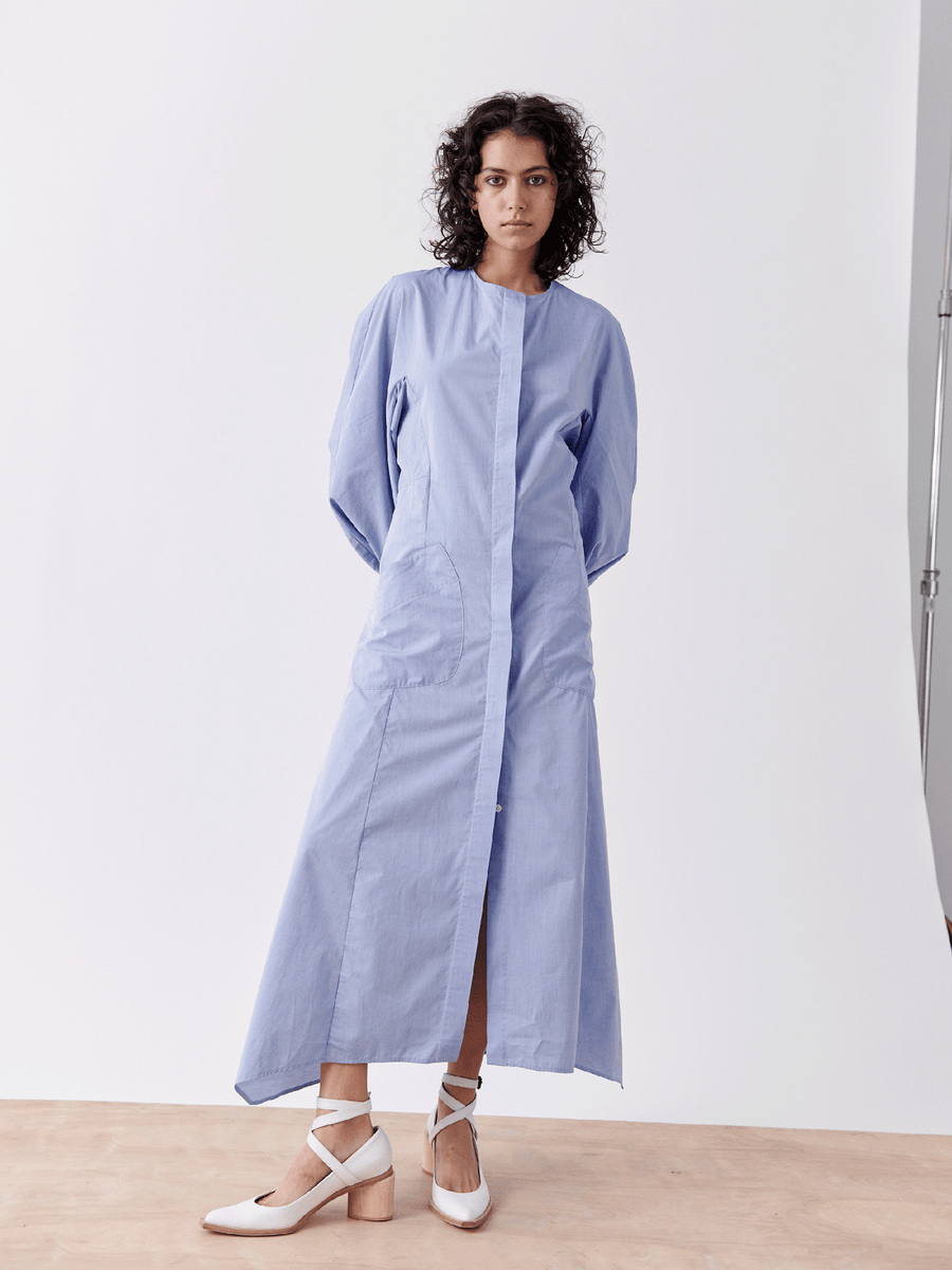 A person with curly hair stands against a white background, wearing the Pleat Akeo Shirt Dress by Zero + Maria Cornejo. Their hands are behind their back, and they complement the look with stylish white block-heeled shoes.