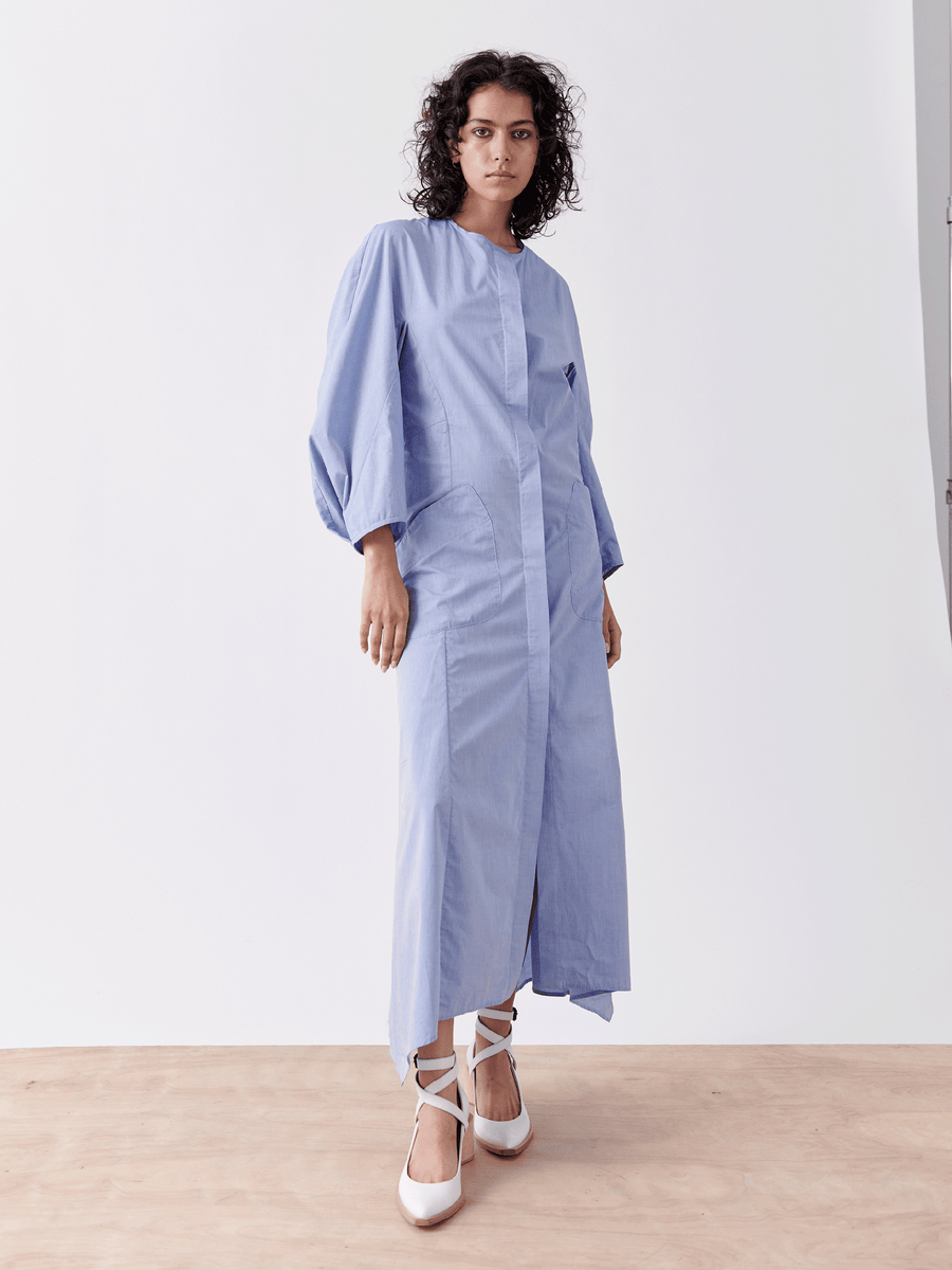 A person with curly hair is wearing the Pleat Akeo Shirt Dress, a light blue chambray piece by Zero + Maria Cornejo, featuring balloon sleeves. They pair it with white ballet-style shoes, standing against a plain white background on a light wooden floor.