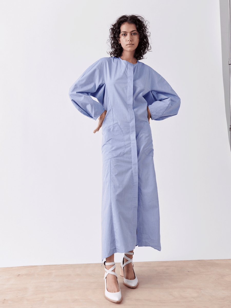 A curly-haired individual models the Pleat Akeo Shirt Dress by Zero + Maria Cornejo. Made of chambray fabric, it features balloon sleeves and a front closure. They stand on a wooden floor against a white background, wearing white shoes, embodying a light and breezy summer look.