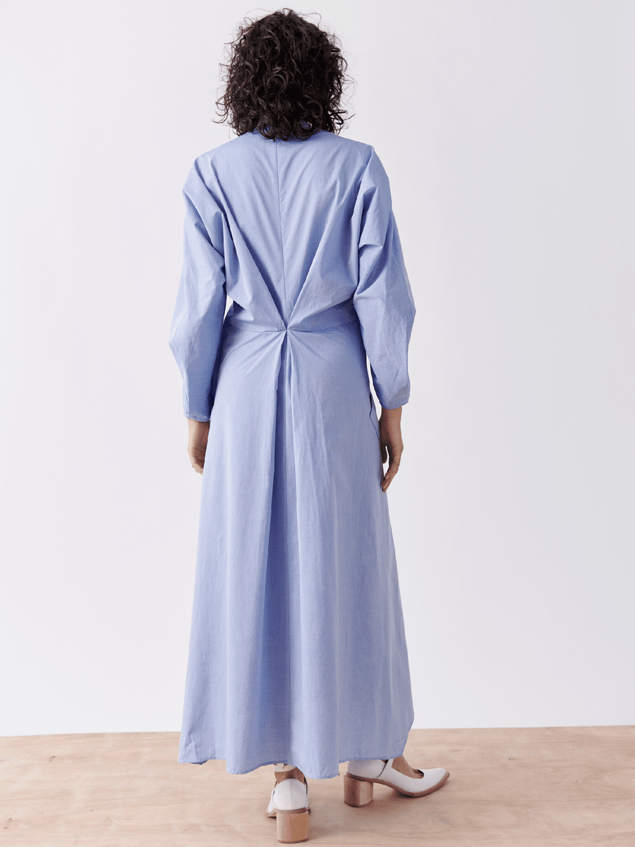 A person with curly hair stands on a wooden floor facing away, wearing the Zero + Maria Cornejo Pleat Akeo Shirt Dress – Final Sale, a long-sleeved light blue cotton chambray dress featuring a pleated back. The look is completed with white shoes.