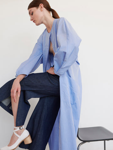 A person with brown hair tied back is seated on a black chair, posing with one knee raised. They wear the Pleat Akeo Shirt Dress by Zero + Maria Cornejo and dark denim pants, paired with white shoes featuring ankle straps against a plain white wall.