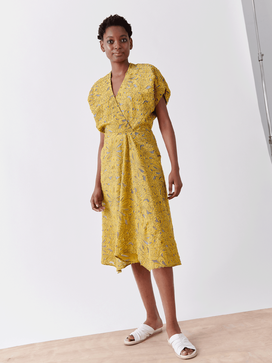 Wearing the Zero + Maria Cornejo Aki Wave Dress – Final Sale, a person stands confidently in yellow. Crafted from organic cotton yarns, it's paired with white sandals against a minimalist studio backdrop for an airy feel.