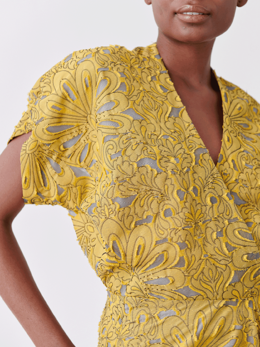 A person in the Aki Wave Dress from Zero + Maria Cornejo, featuring a yellow floral textured design made of organic cotton with short sleeves and a V-neckline, showcasing intricate patterns on a light neutral background.