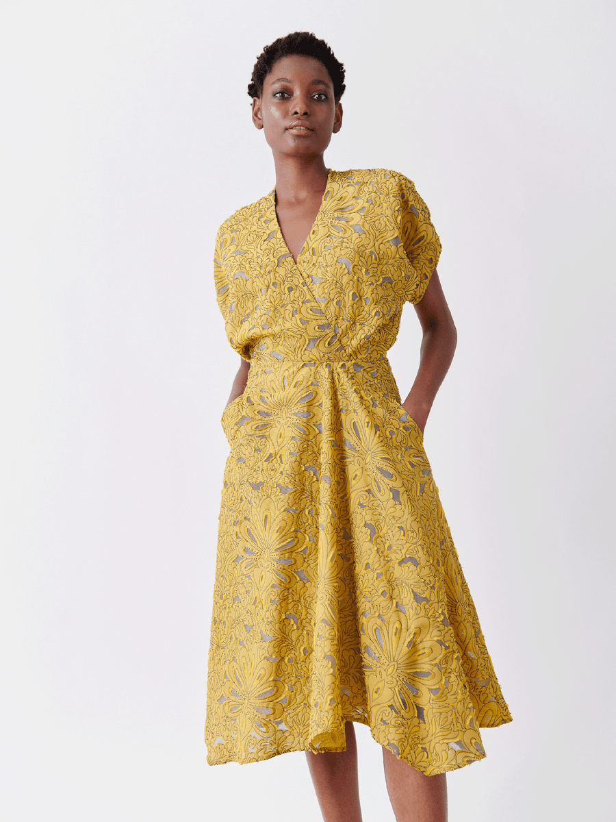 A confident short-haired person models the Aki Wave Dress by Zero + Maria Cornejo against a white backdrop. The yellow, patterned wrap-effect v-neck dress, crafted from organic cotton yarns, features a floral design and short sleeves with pockets.