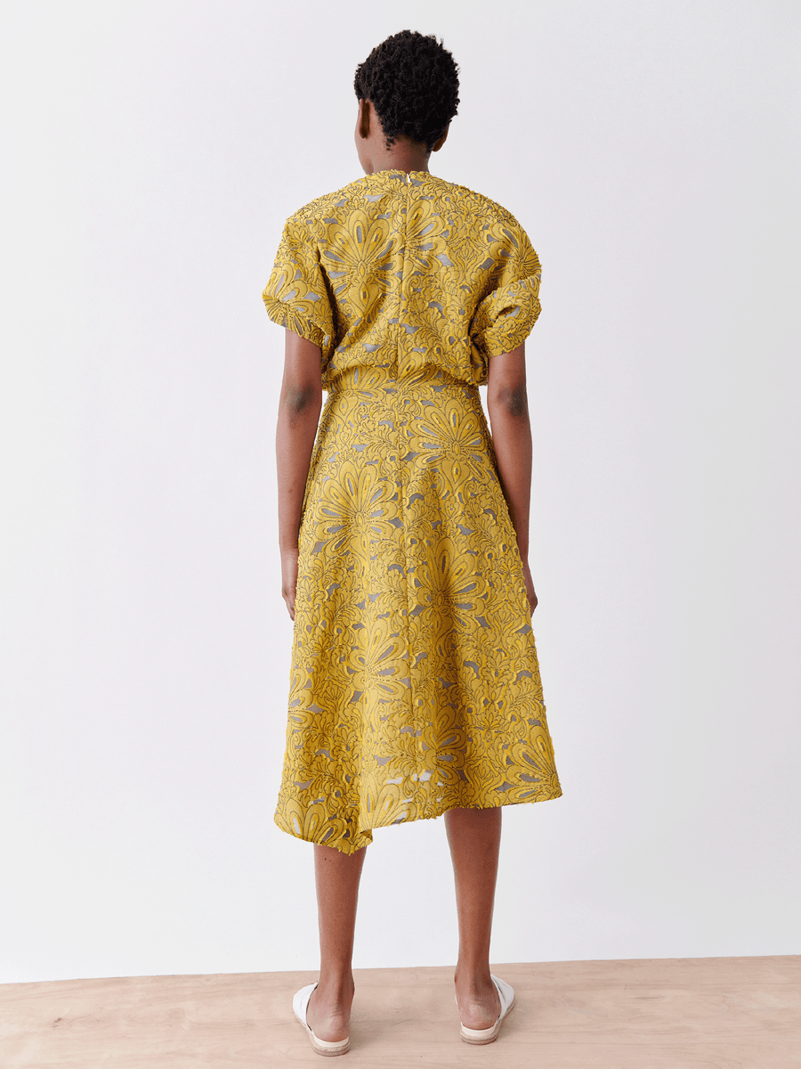 Someone in an Aki Wave Dress by Zero + Maria Cornejo, showcasing a yellow patterned wrap-effect V-neck with short sleeves and knee-length skirt, stands facing away on a light-colored floor against a plain white wall. The dress is crafted from organic cotton yarns and is on final sale.