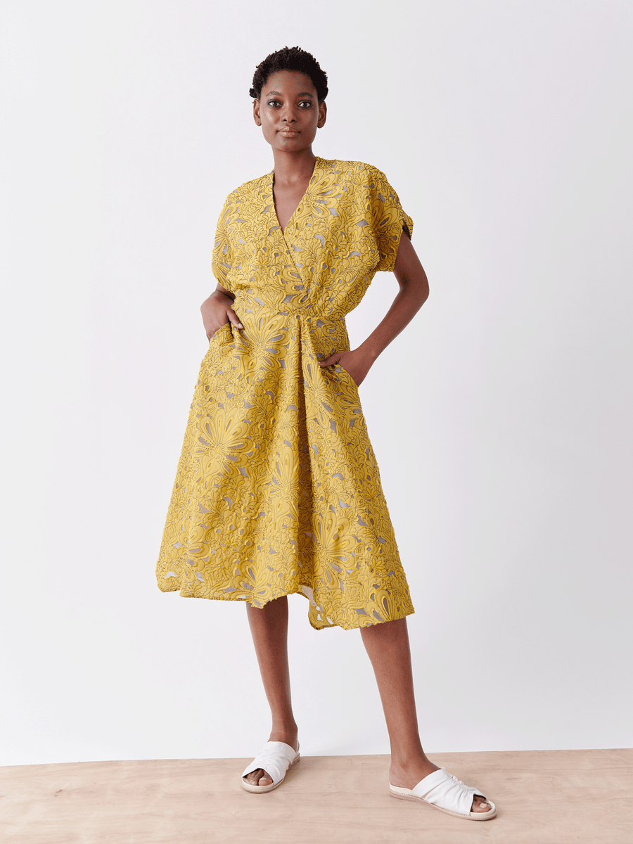 A person confidently stands on a wooden surface, wearing the Aki Wave Dress by Zero + Maria Cornejo, a yellow Damask lace dress with floral patterns and pockets. They pair it with white slip-on sandals, against a plain white background.