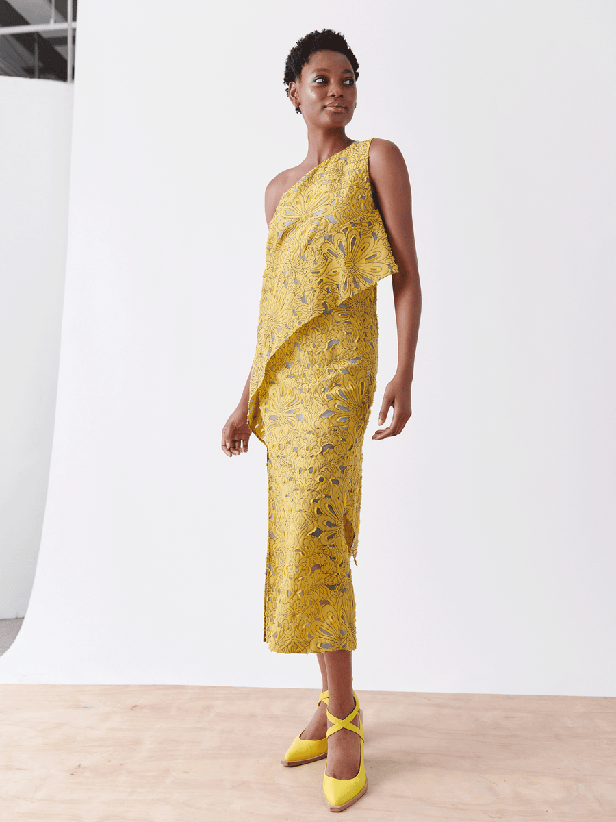 A person in Zero + Maria Cornejo's Long Spiral Dress - Final Sale, a sleeveless yellow sheath with Damask lace jacquard and one-shoulder design, stands on a wooden floor against a white backdrop, wearing matching yellow heels.