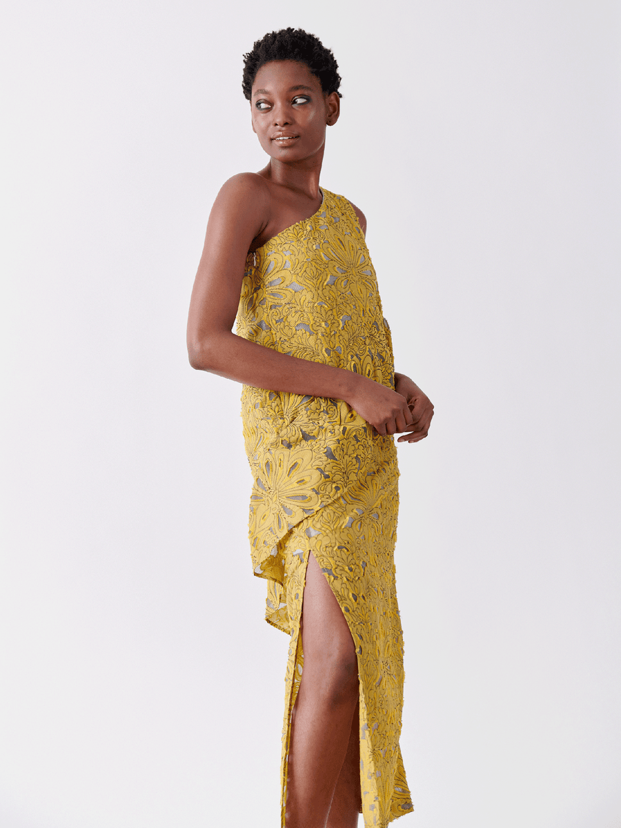 A person in Zero + Maria Cornejo's Long Spiral Dress, a one-shoulder, yellow floral lace sheath dress with a side slit made from organic cotton, poses against a white background. They have short hair and their hands are clasped while gazing to the side.