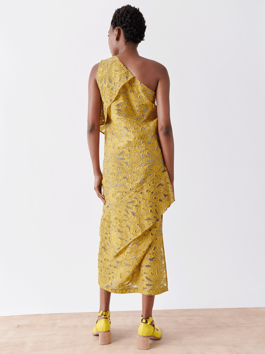 A person stands on a wooden floor, facing away, wearing the Long Spiral Dress by Zero + Maria Cornejo in yellow floral lace. Paired with matching yellow heeled sandals, the organic cotton sheath dress pops against a plain white backdrop.