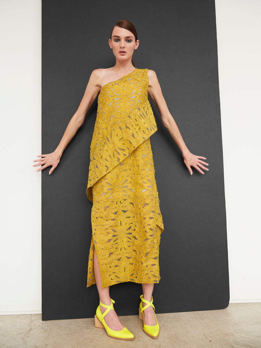 Against a black backdrop, a person showcases the Long Spiral Dress by Zero + Maria Cornejo. This striking one-shoulder, textured yellow dress is crafted from organic cotton with floral patterns and paired with matching shoes, their arms elegantly outstretched.