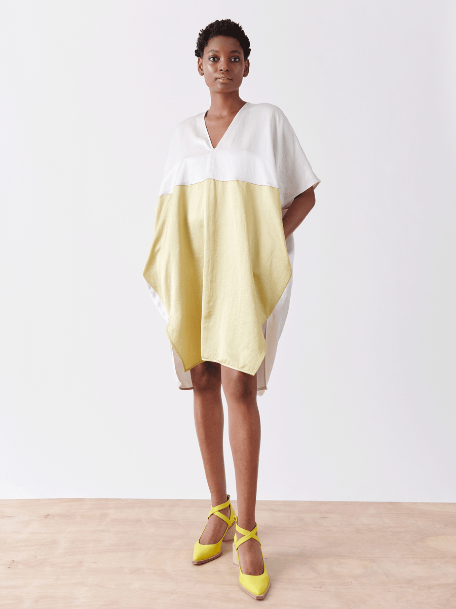 A person stands on a wooden floor wearing the Elie Dress - Final Sale by Zero + Maria Cornejo, a chic linen-blend dress with a white top and light yellow bottom, paired with bright yellow pointed shoes. This New York-made v-neck ensemble is set against a plain white background.