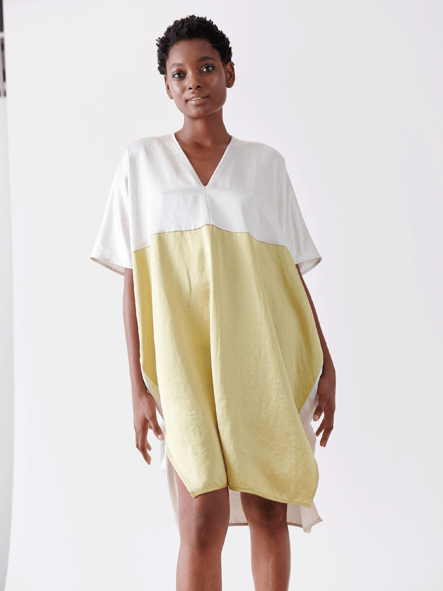 Against a minimalist white backdrop, a person with short hair gently smiles, radiating relaxed confidence in the Elie Dress by Zero + Maria Cornejo. The linen-blend, v-neck outfit features a stylish white top and yellow bottom combination.