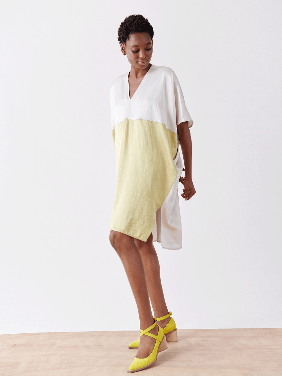 A model in Zero + Maria Cornejo's Elie Dress, a two-tone linen-blend with a white v-neck top and light green draped skirt, pairs it with yellow ankle-strap shoes. She stands against a plain white wall on a light wooden floor.