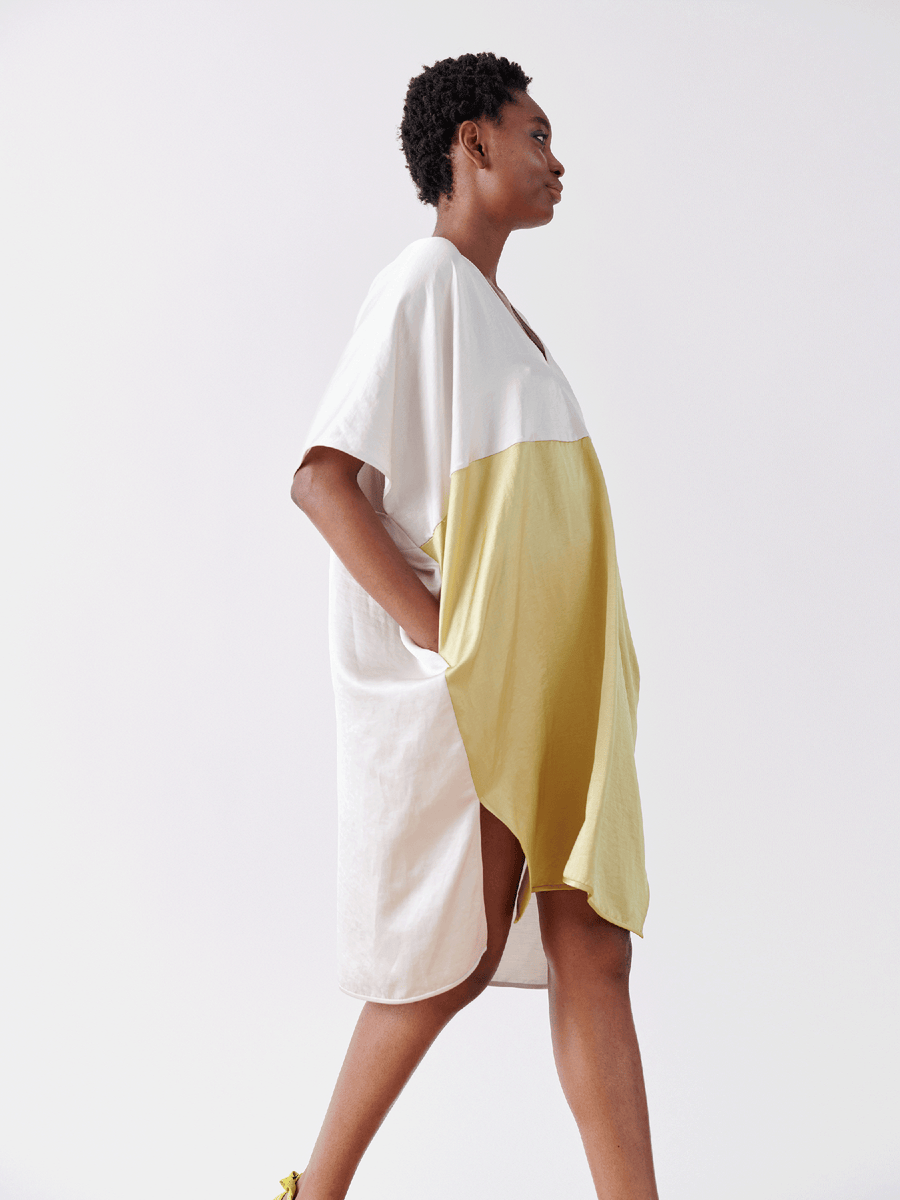 A person in the Elie Dress by Zero + Maria Cornejo, a loose-fitting linen-blend with white and yellow color blocks, short sleeves, a v-neck, and pockets, walks against a plain white background.