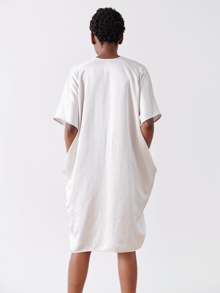 A person, facing away from the camera, stands against a plain background wearing the Elie Dress - Final Sale by Zero + Maria Cornejo. This off-white, linen-blend dress features a draped design with short sleeves and a loose-fitting v-neck style and is made in New York.