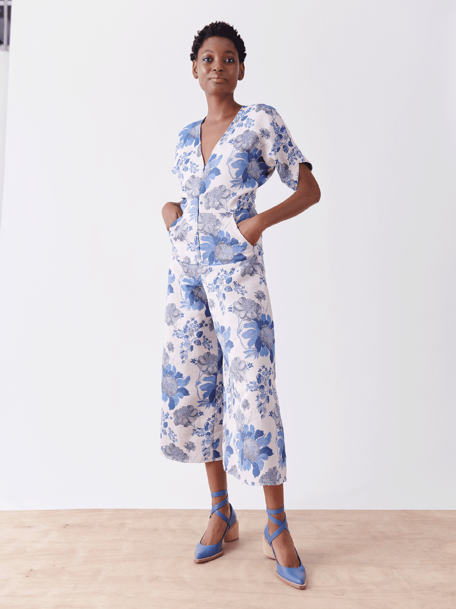 Standing on a wooden floor, a person models the Aissa Eve Jumpsuit by Zero + Maria Cornejo. The white floral jacquard jumpsuit boasts large blue patterns and cropped wide legs, elegantly revealing light blue strappy heels. Hands rest in pockets against a plain white backdrop.