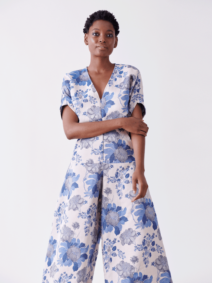 Wearing the Aissa Eve Jumpsuit by Zero + Maria Cornejo, a person stands confidently. This light, floral jacquard outfit features blue and gray blooms on organic cotton with short sleeves and wide-legged pants. The look embodies sustainable fashion against a plain white background.