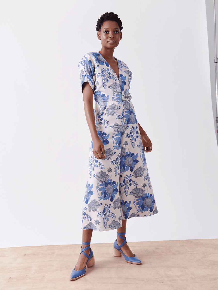 A person poses confidently in the Aissa Eve Jumpsuit by Zero + Maria Cornejo, featuring a blue and white floral jacquard design, cropped wide legs, short hair, and blue ankle-strap shoes, against a plain backdrop with one hand on their hip.