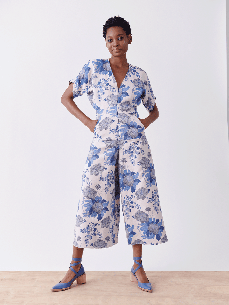 Confidently posed in the Aissa Eve Jumpsuit by Zero + Maria Cornejo, a person with short hair and blue shoes stands gracefully against a white background. The blue and white floral jacquard design features short sleeves and cropped wide-leg pants, making a bold statement.