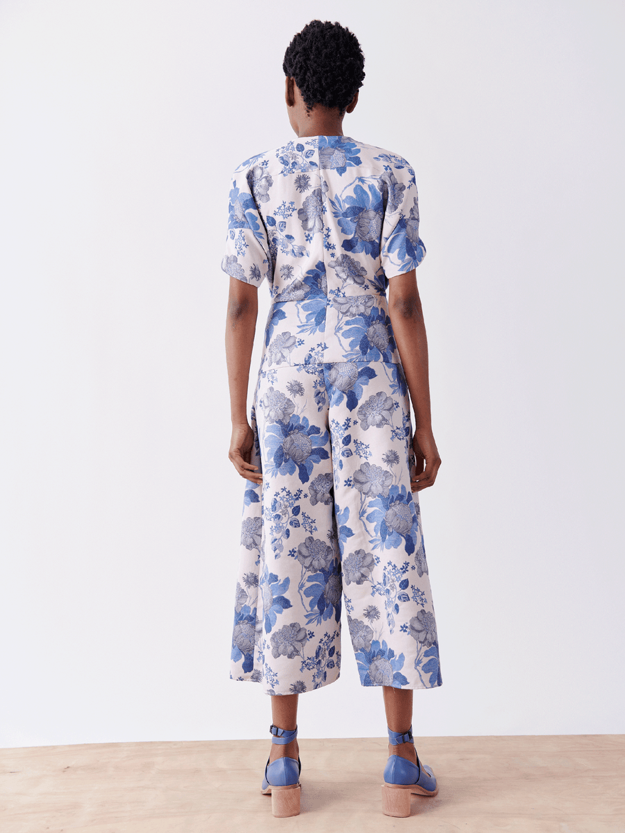 Against a plain white backdrop, an individual stands on a wooden floor wearing the Aissa Eve Jumpsuit from Zero + Maria Cornejo. The blue and white floral jacquard outfit features cropped wide legs and is paired with matching shoes, while their short curly hair enhances the chic look.