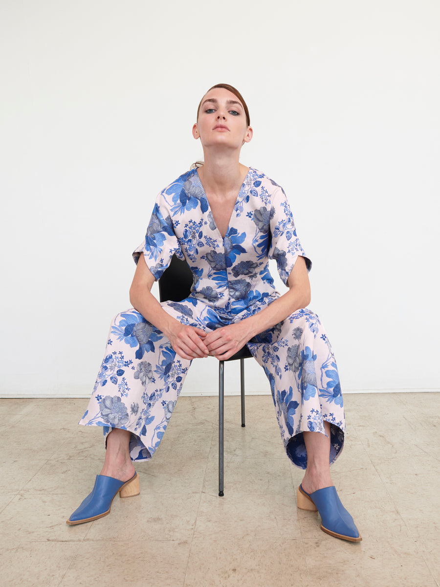 Seated against a plain white wall, a person wears the Aissa Eve Jumpsuit by Zero + Maria Cornejo, featuring a blue and white floral jacquard design. Their short hair frames their face as they look directly at the camera, with chic blue mules perfectly enhancing the outfit's elegance.