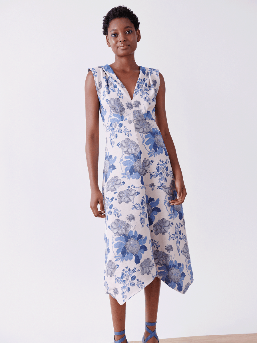 A person confidently poses in the Joi Dress by Zero + Maria Cornejo, a sleeveless V-neck floral jacquard dress made from organic cotton. They stand against a plain white background wearing blue sandals.