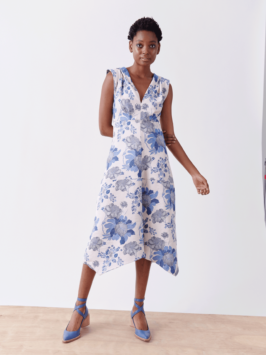 A person confidently models the Joi Dress by Zero + Maria Cornejo on a plain white background. This sleeveless cocktail dress, crafted from organic cotton, features a blue floral pattern, V-neck, and asymmetrical hem. They pair it with matching blue ankle-tie shoes. Final Sale item.